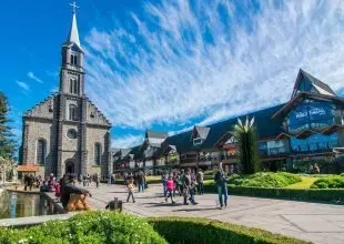 Igreja de pedra e edifício moderno de vidro em uma praça ensolarada com pessoas caminhando e paisagismo verde.