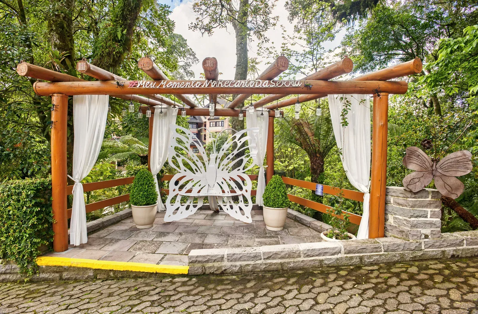 Entrada para eventos com pérgola de madeira, cortinas brancas e decoração floral em um caminho de pedra ao ar livre.