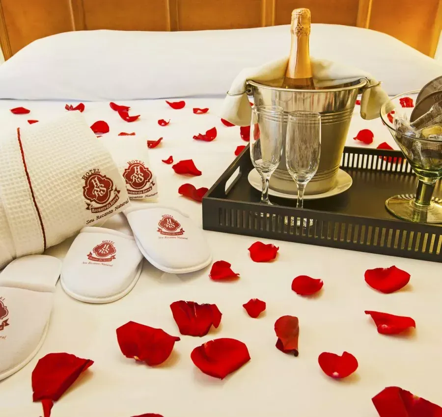 Pétalas de rosa e champanhe sobre uma cama com chinelos e toalhas brancas em um cenário romântico de hotel.