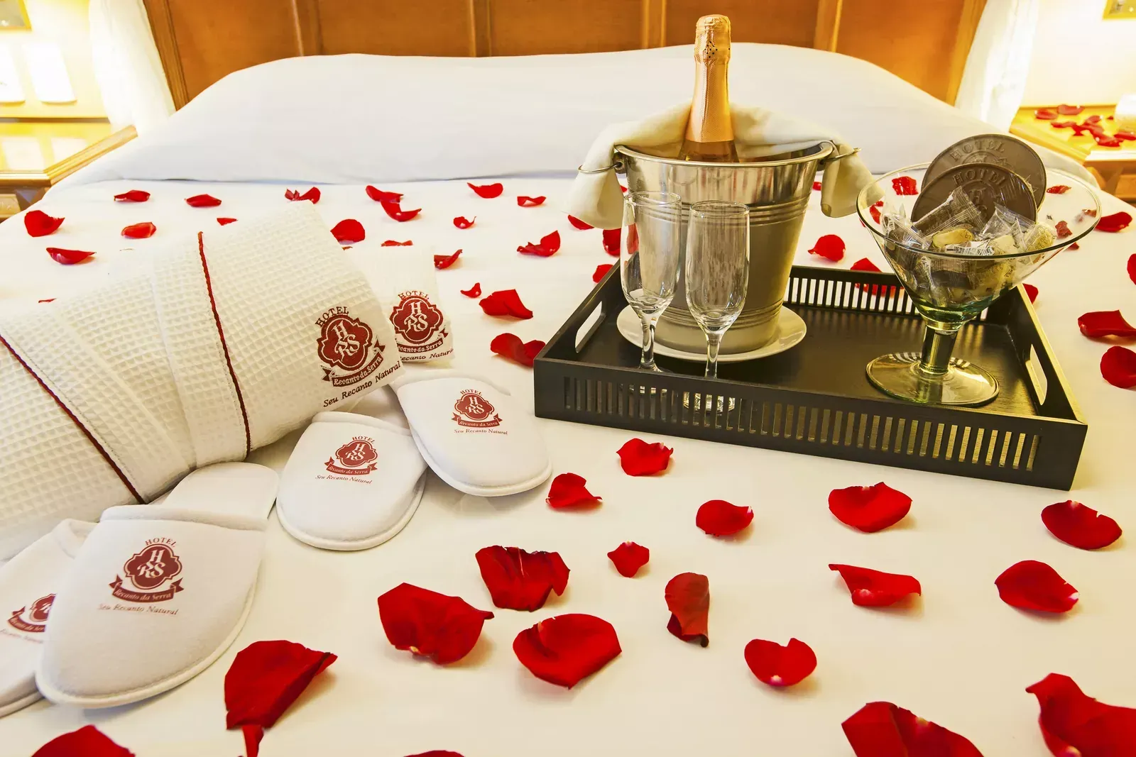 Cama de hotel romântica com pétalas de rosa, toalhas dobradas e um balde de champanhe em uma bandeja.