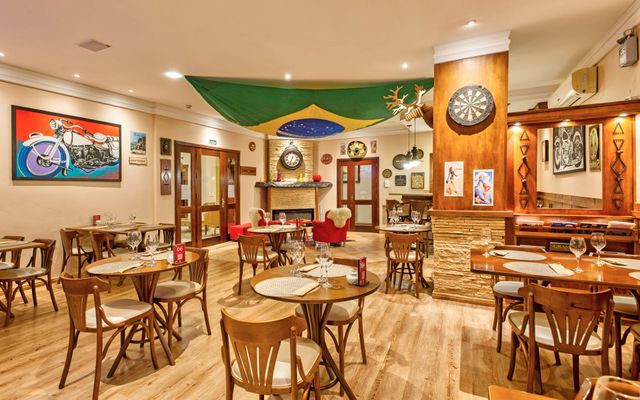 Interior de um restaurante decorado com uma bandeira brasileira, bar, mesas e poltronas vermelhas.