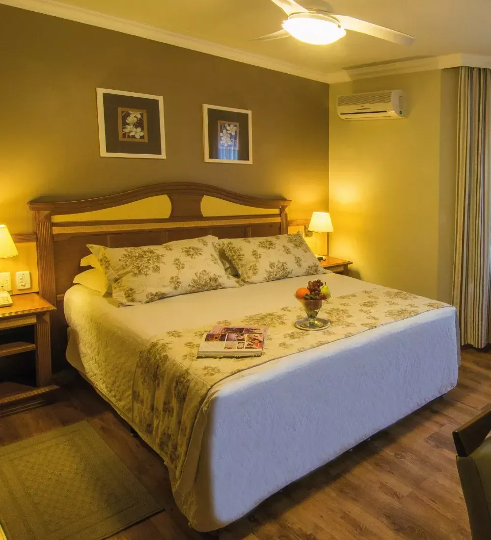Quarto de hotel aconchegante com cama king-size, luminárias com luz quente, obras de arte emolduradas e cortinas na janela.