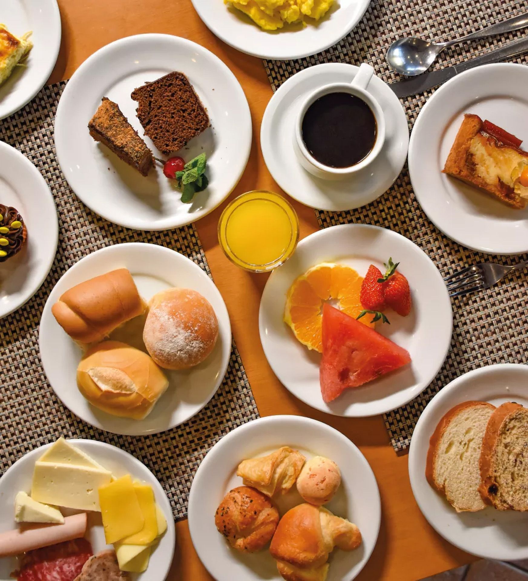 Doces variados e frutas em pratos brancos ao redor de uma xícara de café sobre um jogo americano de palha.