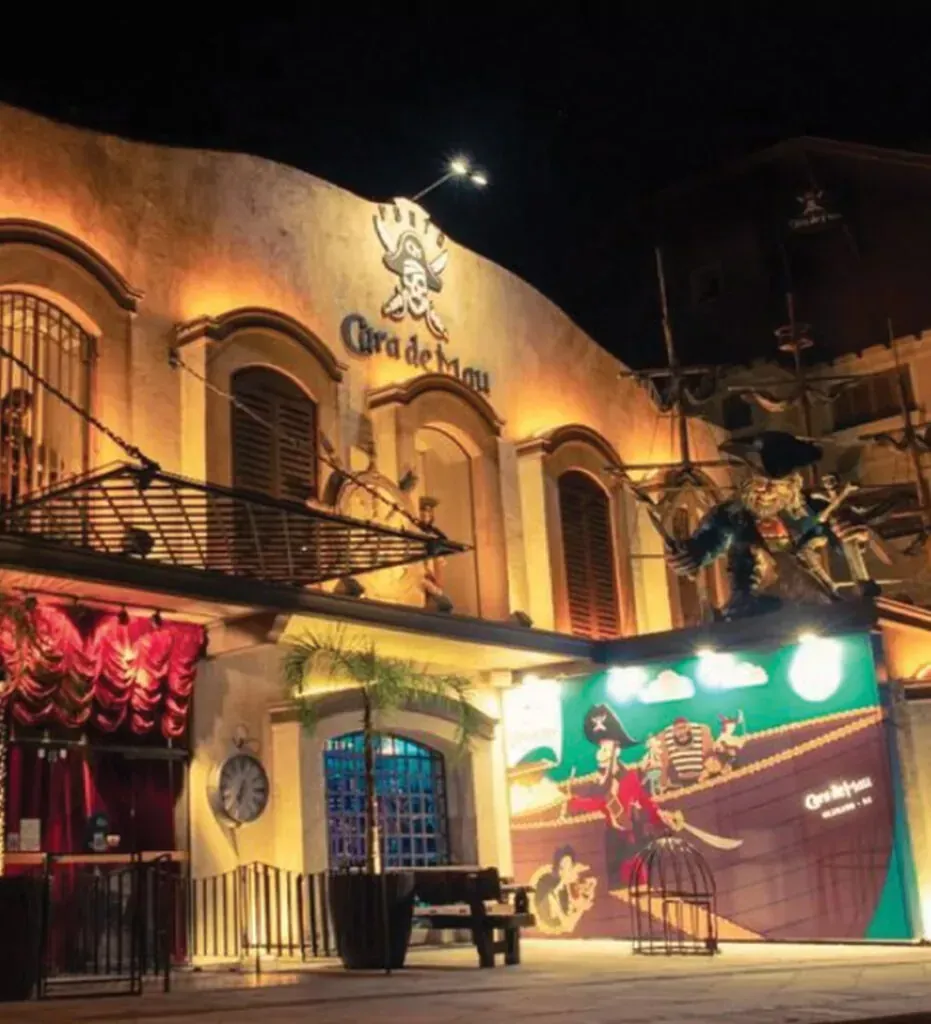 Fachada de loja com iluminação aconchegante, janelas em arco, cortinas vermelhas e um mural colorido à noite.