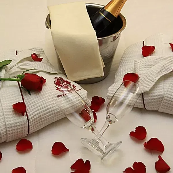 Garrafa de champanhe em balde com guardanapos brancos, pétalas de rosa vermelha e duas taças sobre uma mesa.