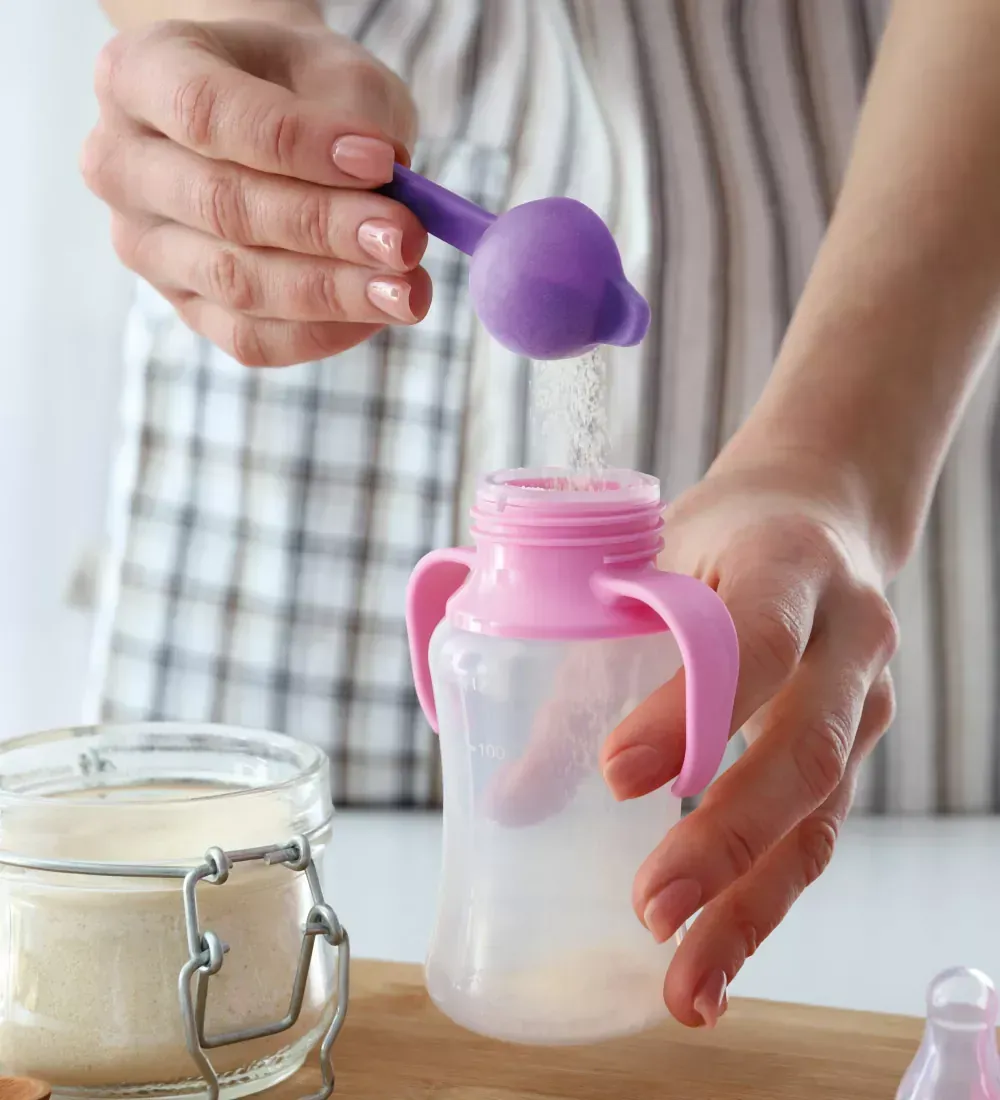 Mãos preparando uma mamadeira com uma colher roxa em uma bancada de cozinha