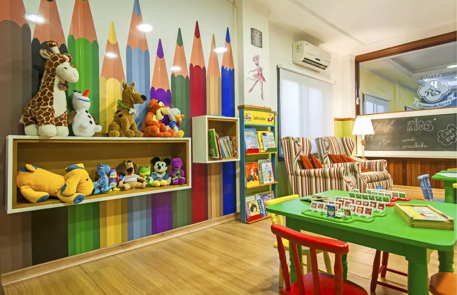 Sala de aula infantil colorida com prateleiras de brinquedos, lápis e uma mesa de atividades verde.