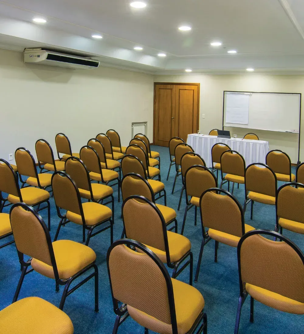 Sala de conferências vazia com fileiras de cadeiras amarelas, um quadro branco e uma mesa na frente.
