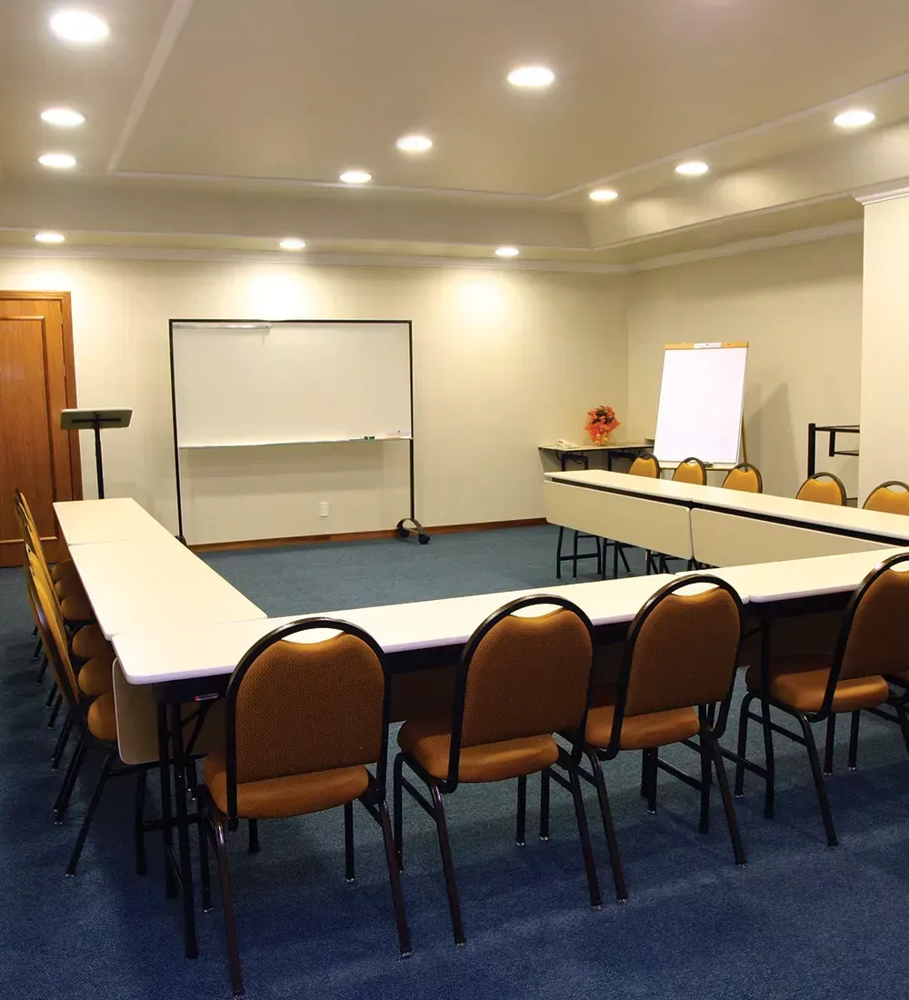 Sala de conferências vazia com mesas em formato de U, cadeiras, quadro branco e tela de apresentação sobre carpete azul.