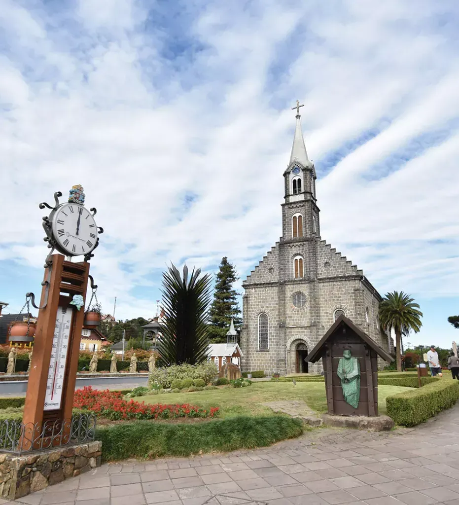 Igreja de pedra com torre sineira em um jardim paisagístico sob um céu nublado, ao lado de um relógio decorativo.