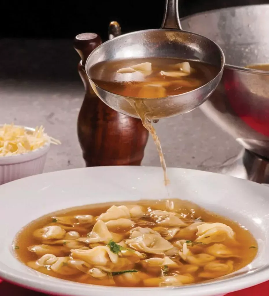 Concha despejando caldo em uma tigela de sopa de bolinhos sobre uma mesa com condimentos por perto.