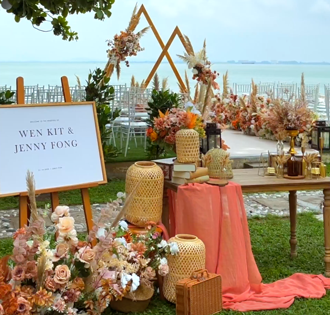 A wedding setup on a lawn by the sea featuring a welcome sign, boho floral arrangements, and wooden lanterns.