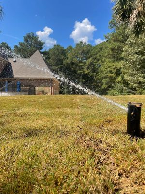 A lawn sprinkler spraying water across a dry, grassy yard toward a house under a clear, blue sky.