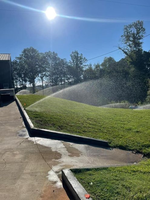 A sprinkler waters a green grassy hill under a bright sun, with a concrete walkway in the foreground.
