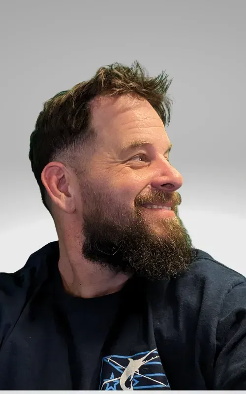 Side profile of a smiling person with a dark, full beard, wearing a dark blue shirt with a graphic design.