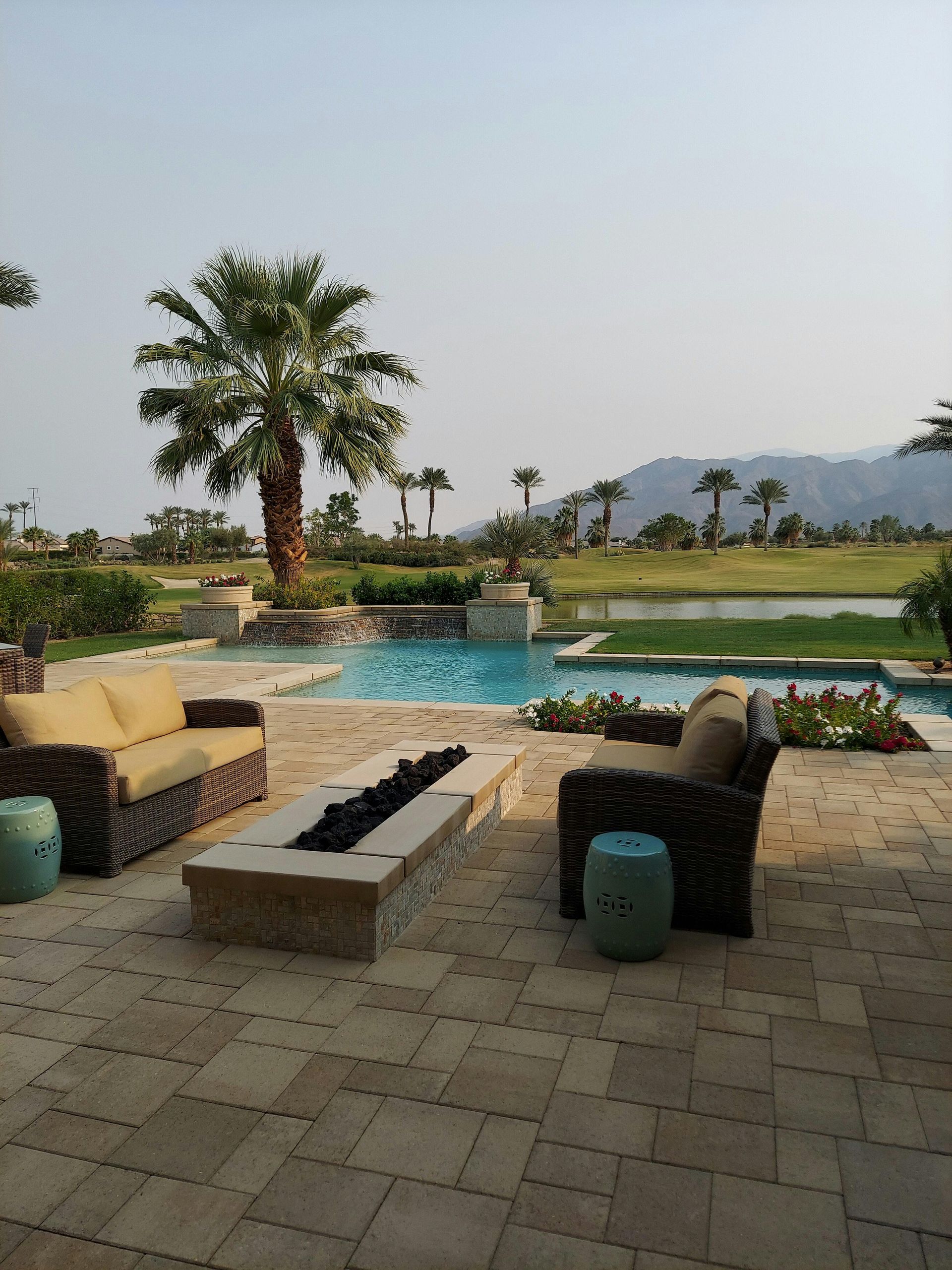 A patio with two wicker sofas and a fire pit overlooking a swimming pool, lawn, palm trees, and distant mountains.