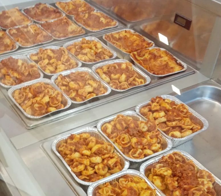Piatti di pasta preparati in vassoi di alluminio, esposti in un banco gastronomia refrigerato.