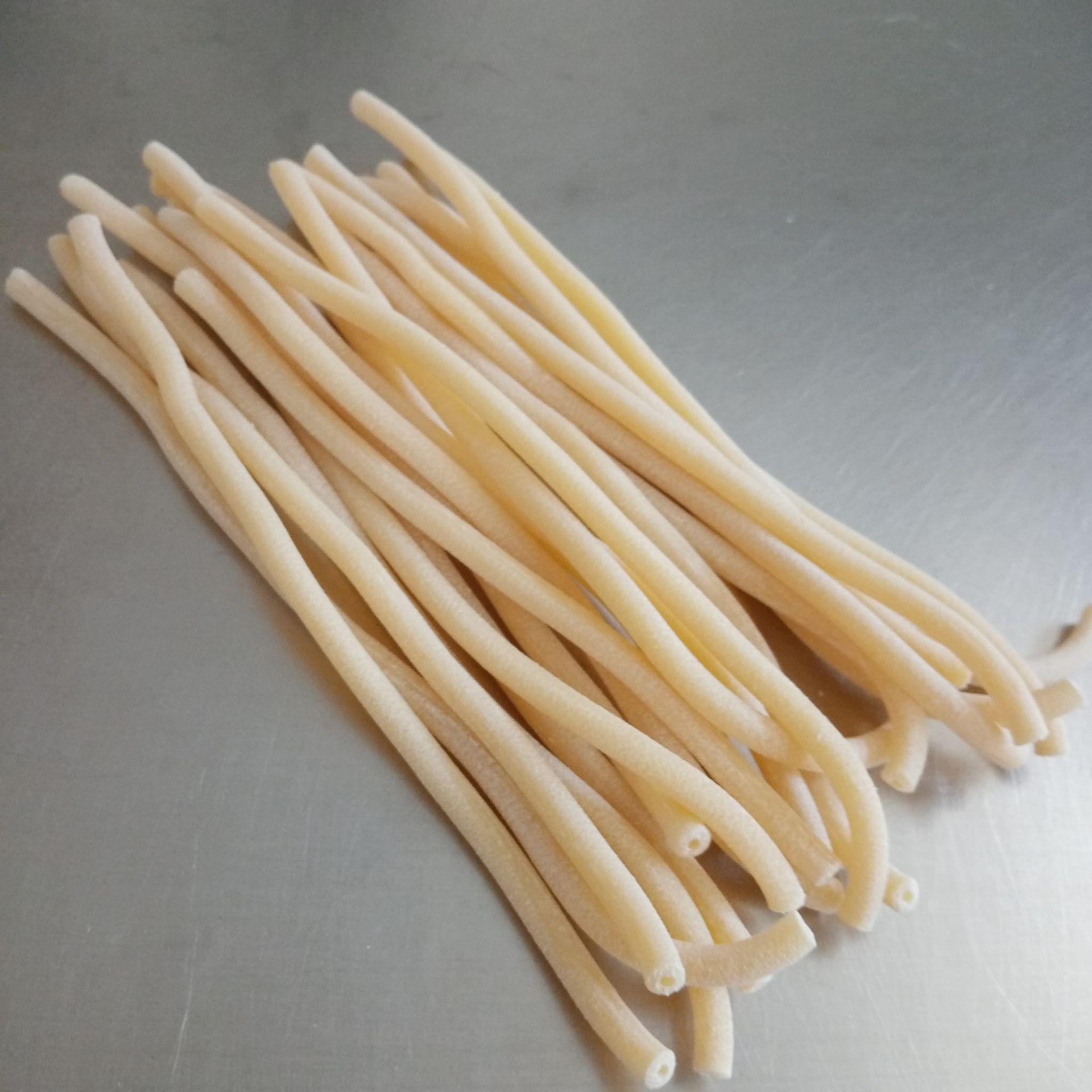 Mucchio di tagliatelle di pasta tubolari, spesse e di colore chiaro, su una superficie metallica.