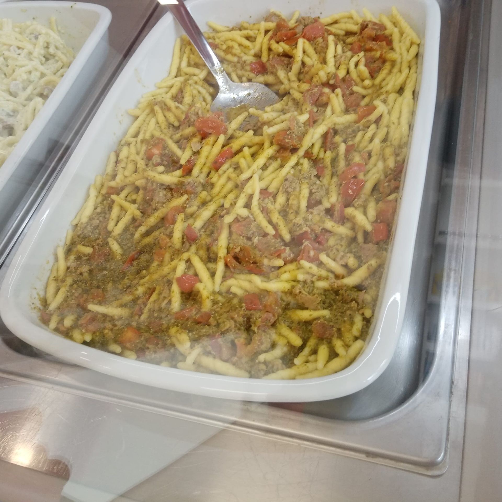 Piatto di pasta in un vassoio bianco con pomodori e condimenti, cucchiaio al centro.