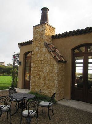 Stone patio beside a rustic stucco building with arched windows, outdoor table and chairs, and a chimney.