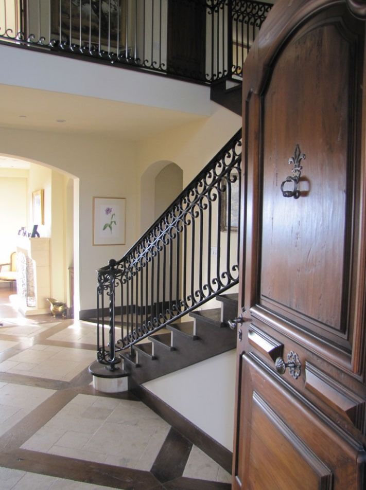 Elegant foyer with curved staircase, ornate railing, and large wooden doors; sunlight on tiled floor