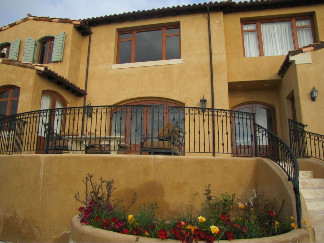 Two-story stucco house with arched windows, black railing, and a flower bed in front