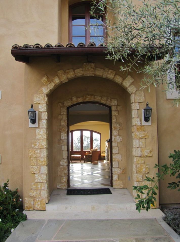 Stone archway entrance to a home with a wooden door, lanterns, and a courtyard view beyond.