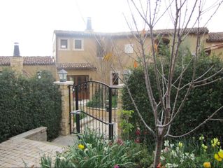 Front garden with wrought-iron gate, flowering plants, and a beige house behind trees