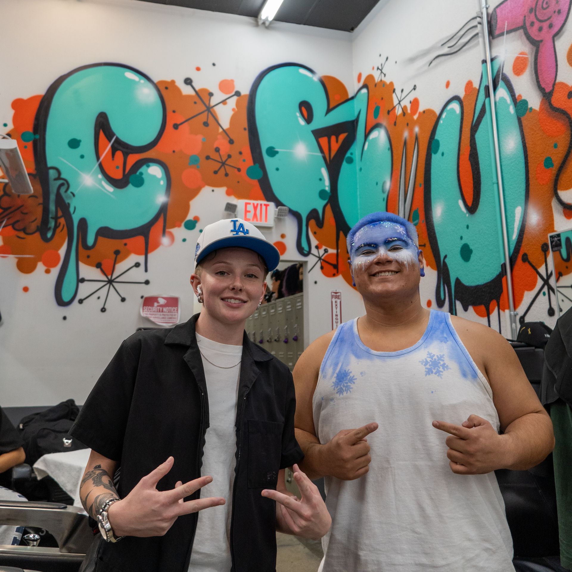 Two people posing in front of colorful graffiti art, smiling and making hand signs.