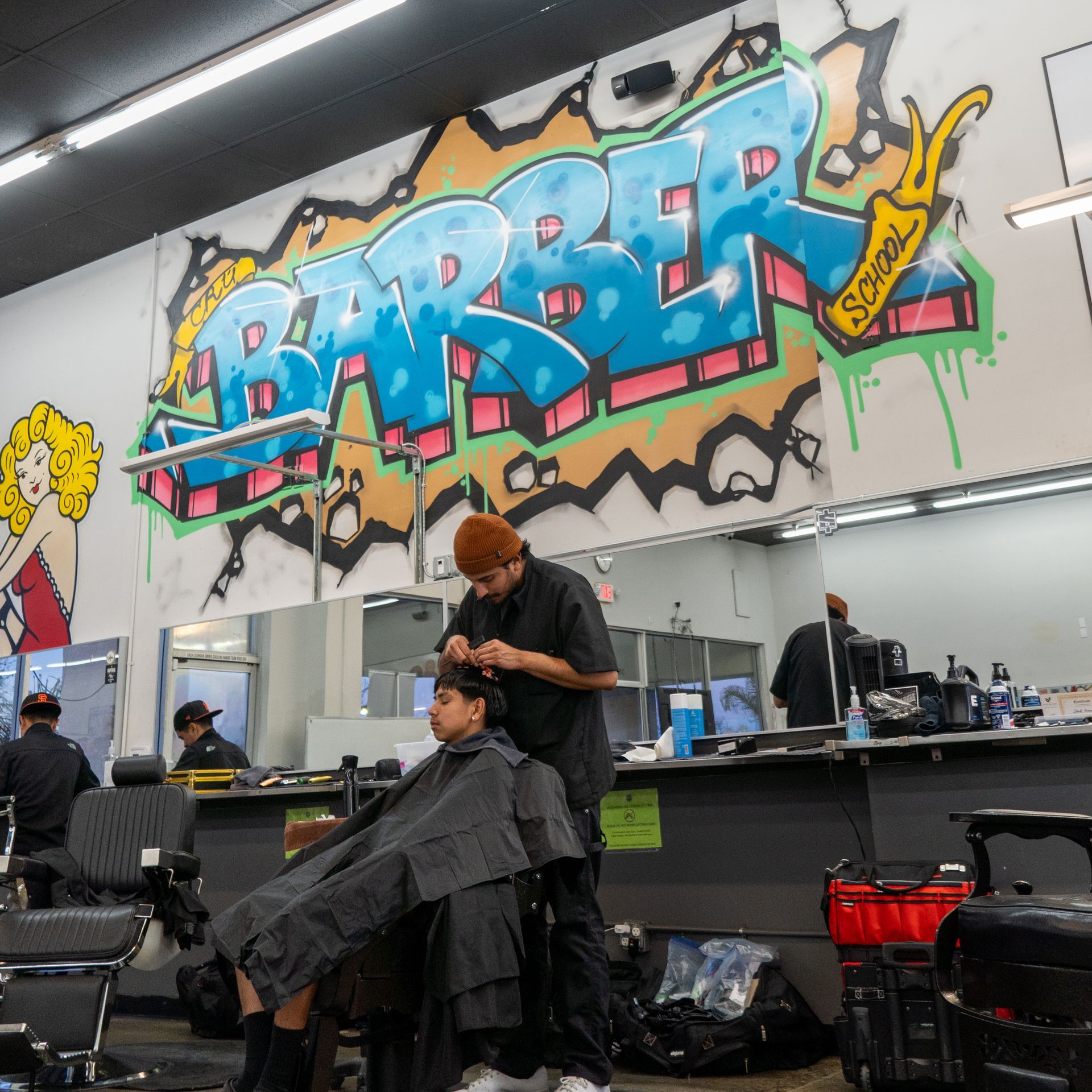 Barber cutting a client’s hair in a shop with a large colorful graffiti mural on the wall
