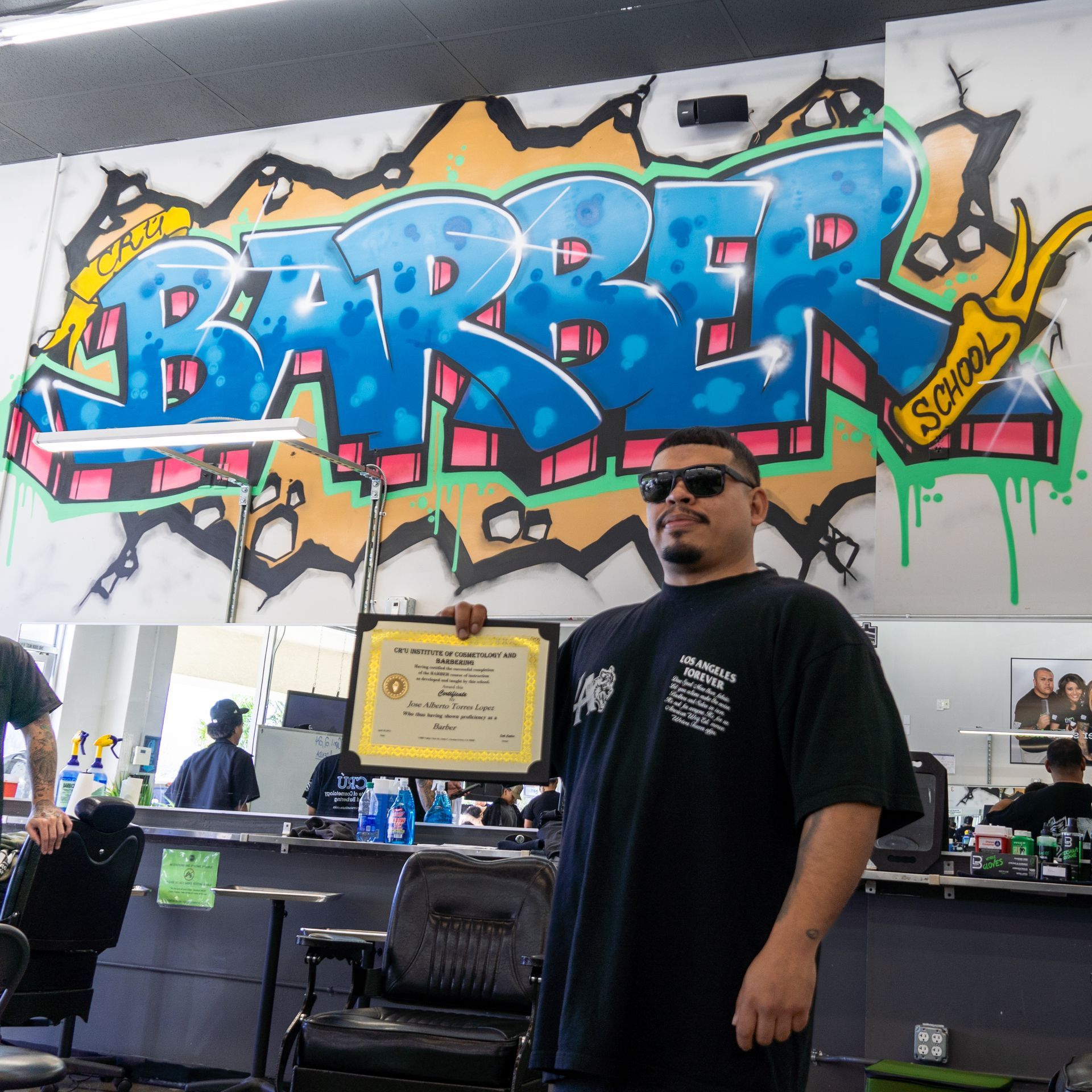 Man standing in a barber shop beneath colorful graffiti reading “BARBER,” holding a framed certificate.