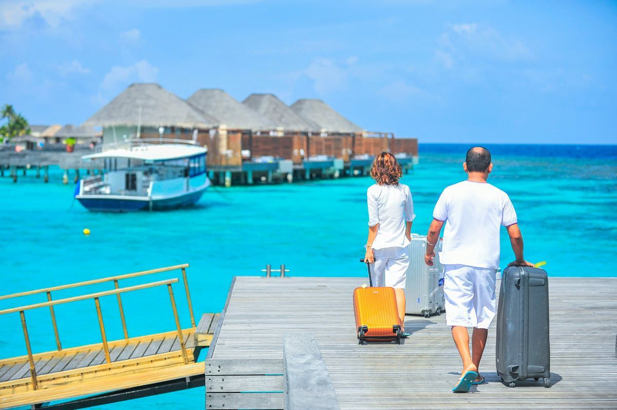 Two travelers walk on a wooden pier with suitcases toward overwater bungalows in turquoise water.