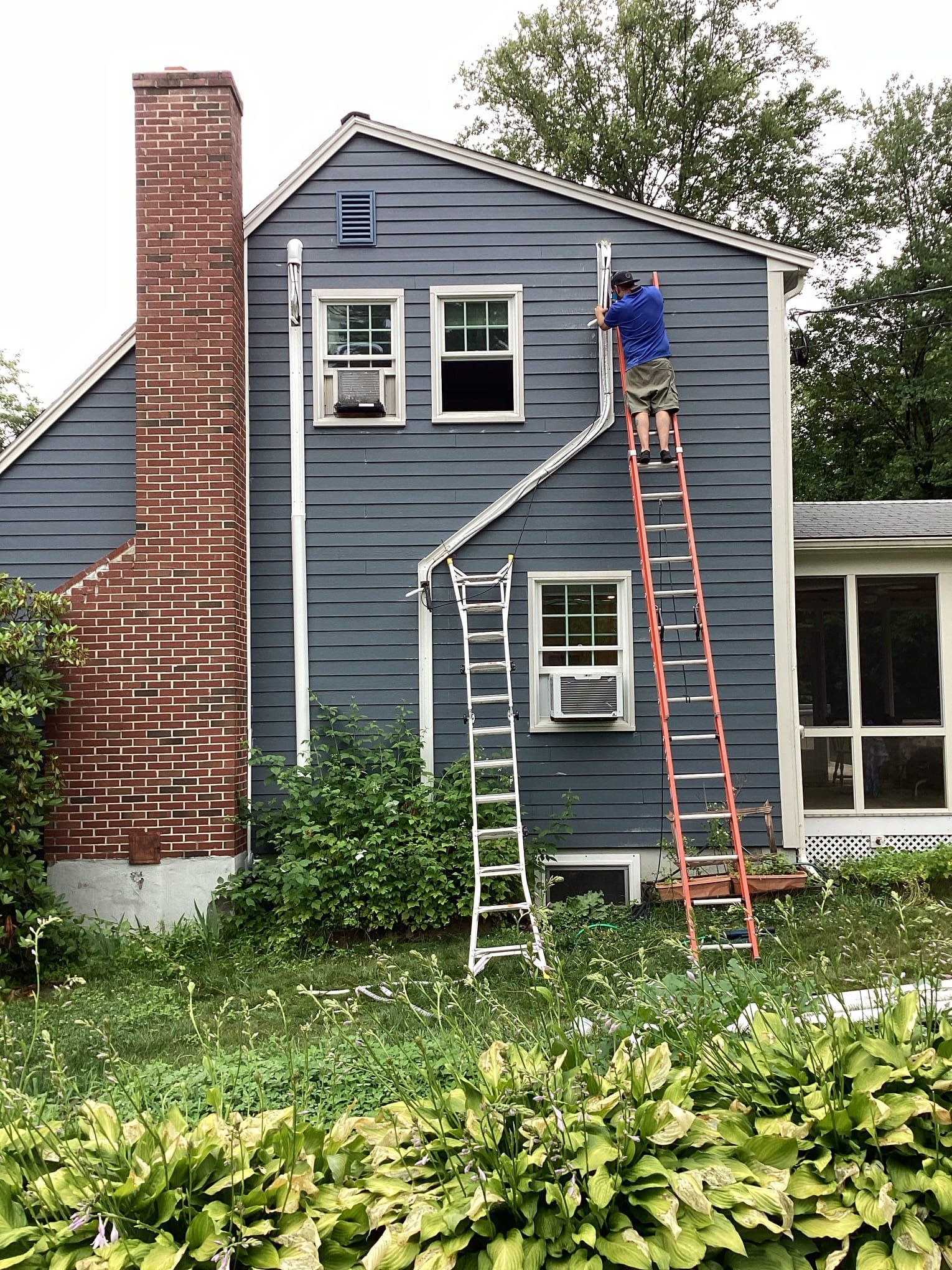 Person on ladder fixing siding on blue house with brick chimney.