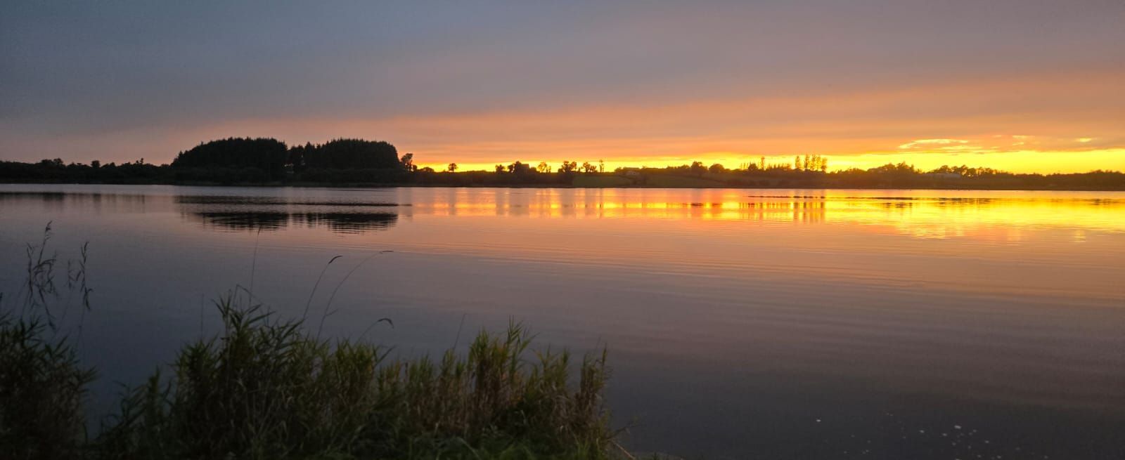 Person relaxing in a hot tub overlooking a calm lake and trees at sunset
