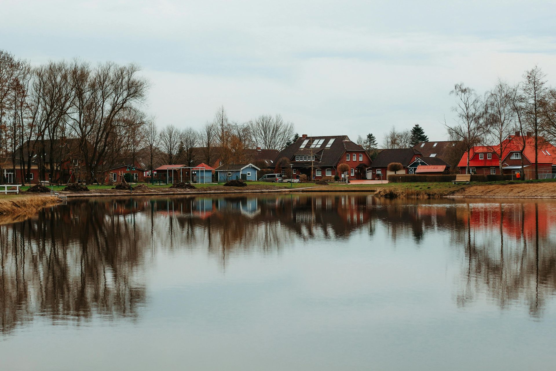 Calm lake reflecting bare trees and colorful houses along the shore under a cloudy sky