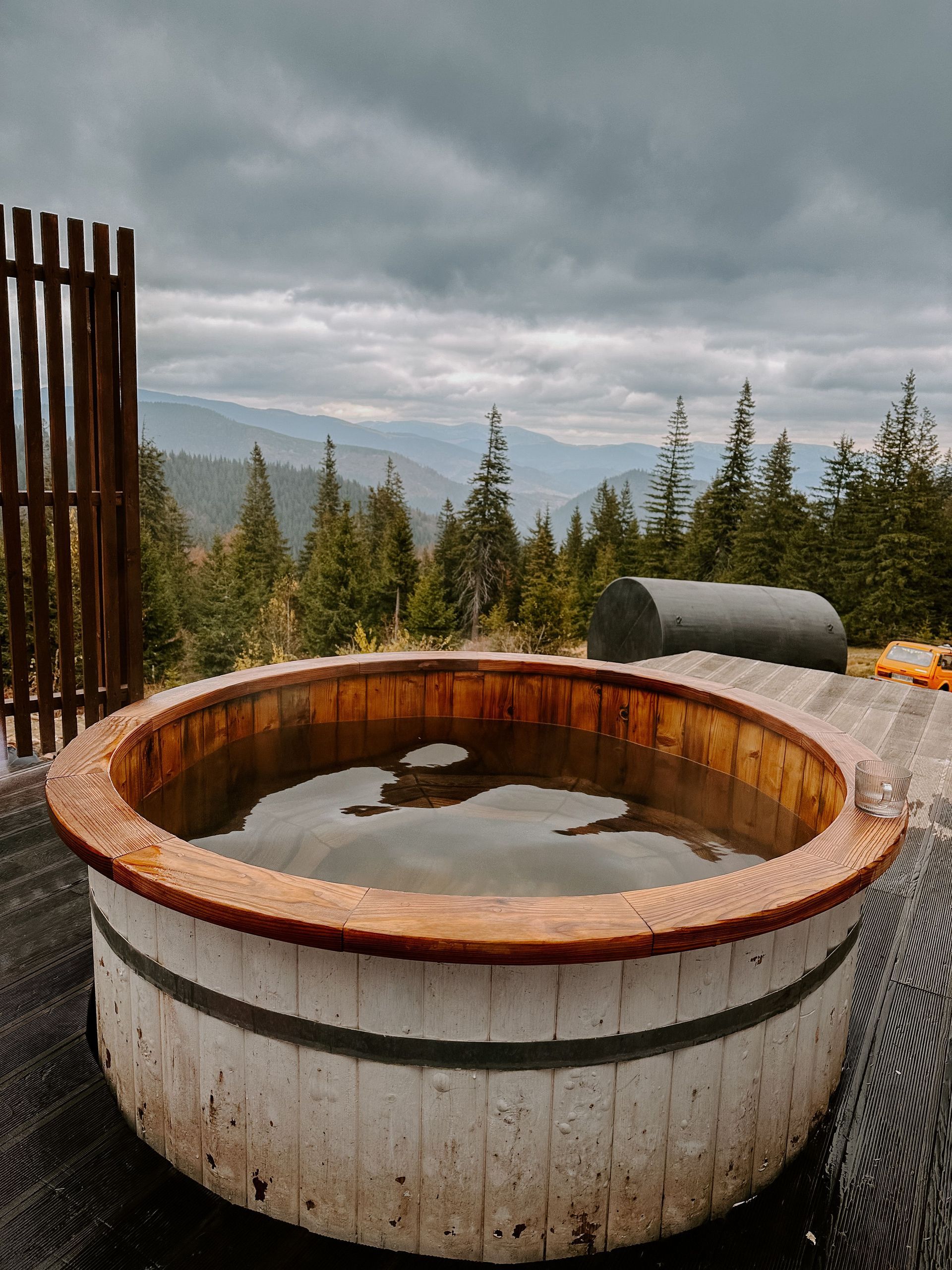 Outdoor wooden hot tub with still water, overlooking pine trees and mountains under a cloudy sky