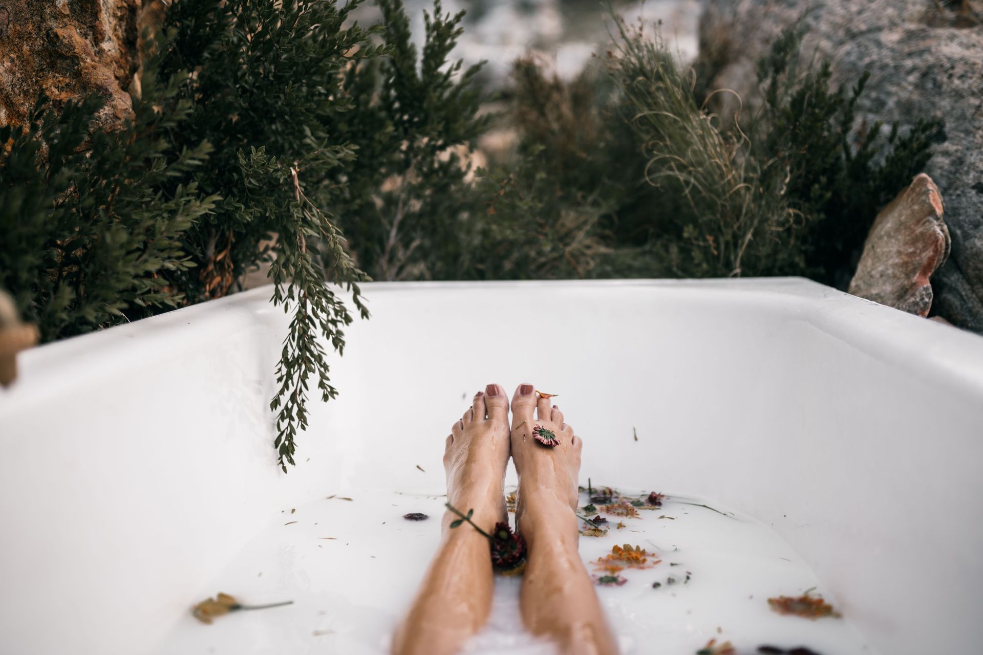 Feet soaking in a white bathtub outdoors, with leafy plants in the background.
