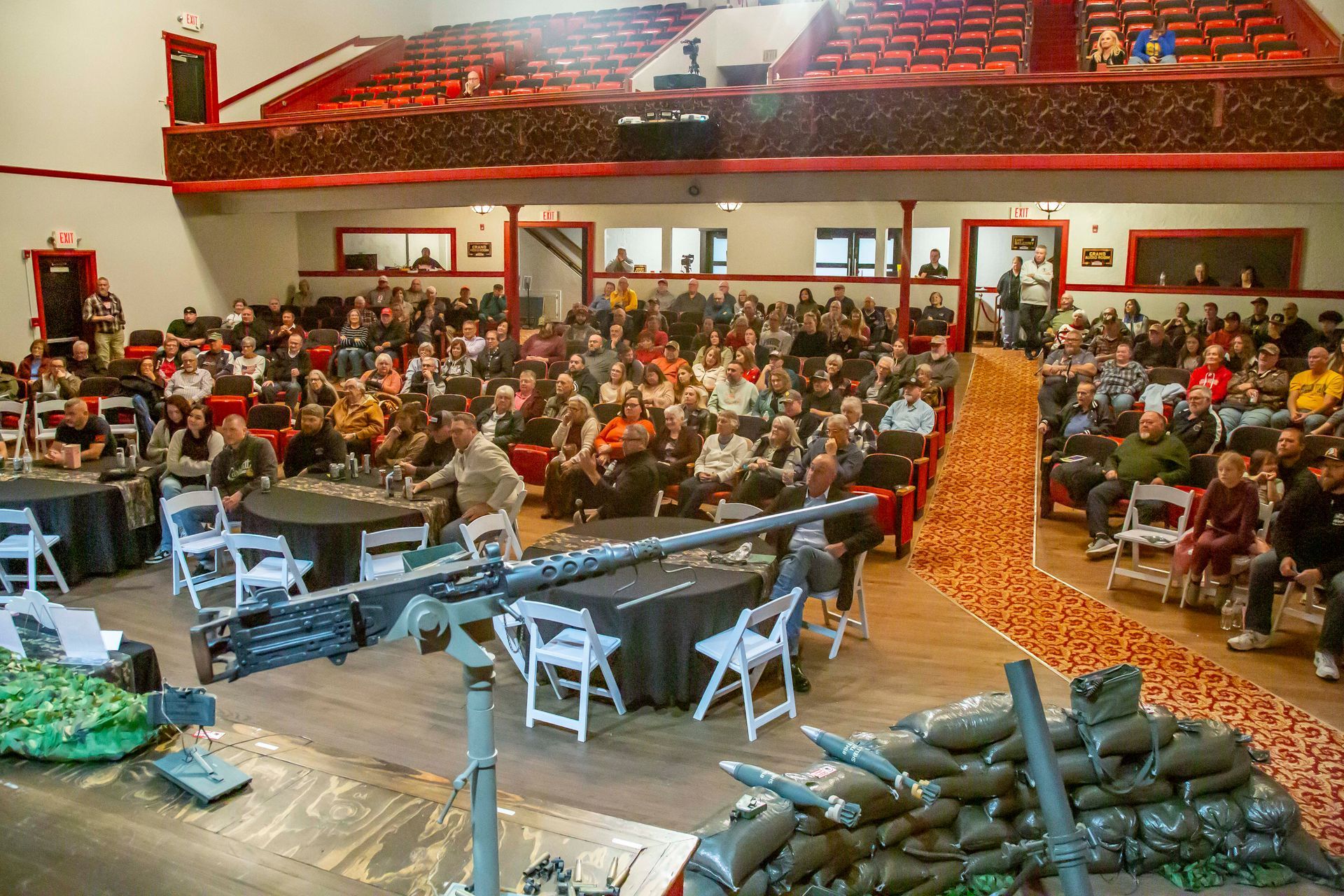 Audience in auditorium with stage set up, gun display, and rows of seating.