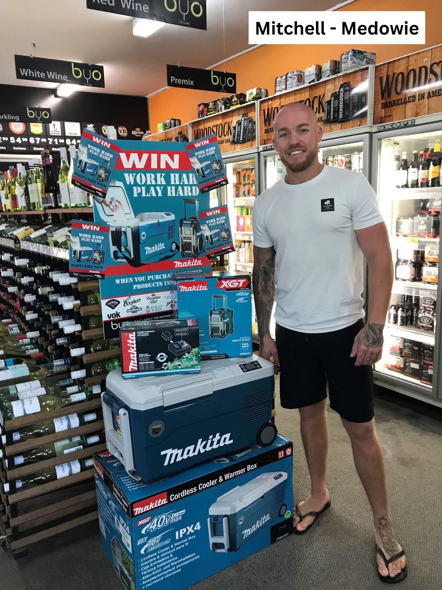 Man stands by stacked Makita products in a store. He smiles, wearing a white shirt and black shorts.