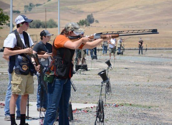 Two people aim shotguns at an outdoor shooting range with desert hills in the background.