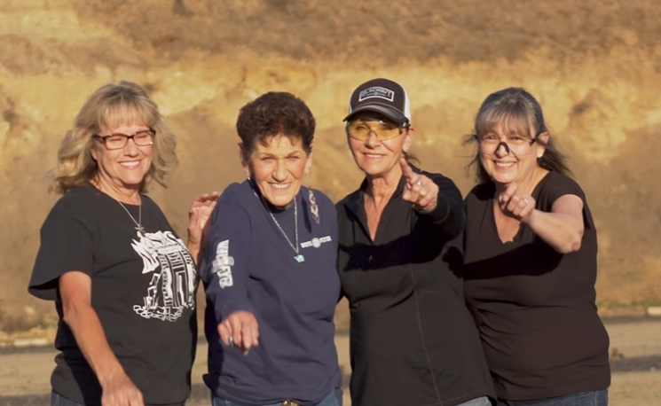 Four people standing outdoors in black shirts, smiling and pointing at the camera.