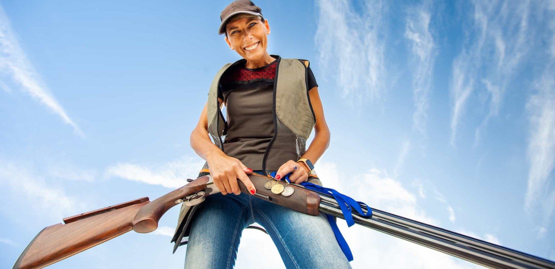 Smiling hunter holding a shotgun, standing outdoors under a bright blue sky
