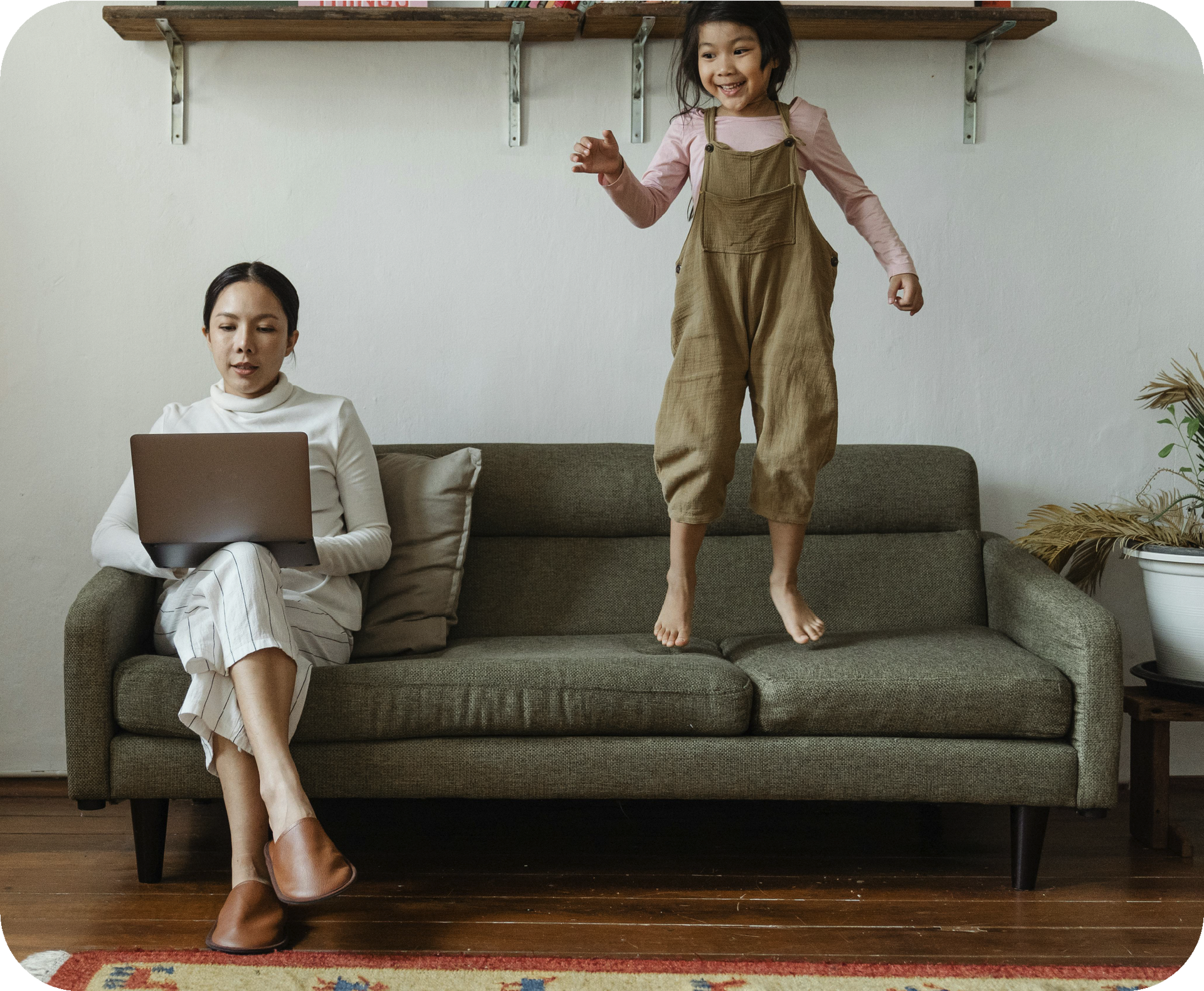 Woman using a laptop on a couch while a child jumps beside her in a living room.