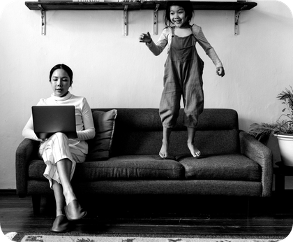 A woman is sitting on a couch with a laptop and a child is jumping on the couch.