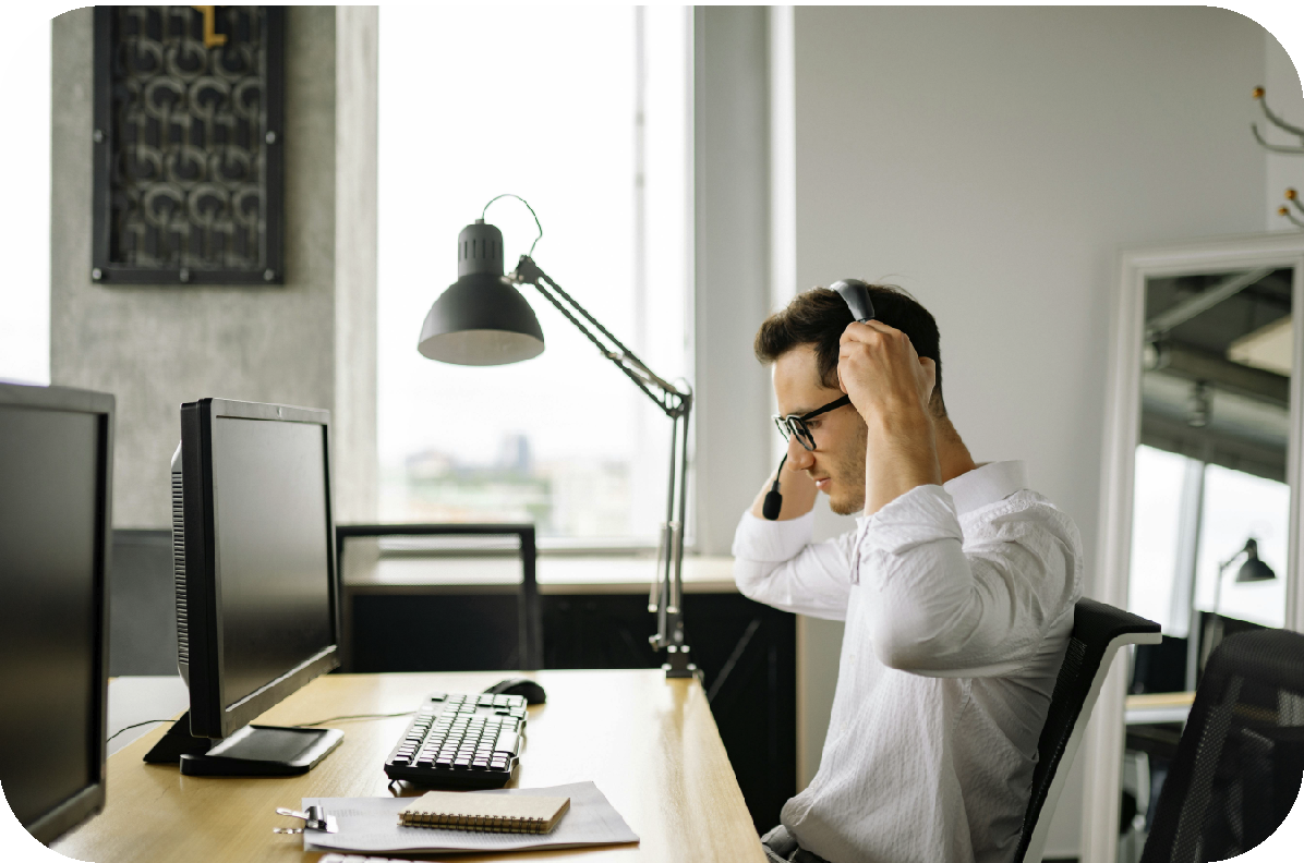 Person at a desk putting on headphones in a bright office with two monitors and a lamp