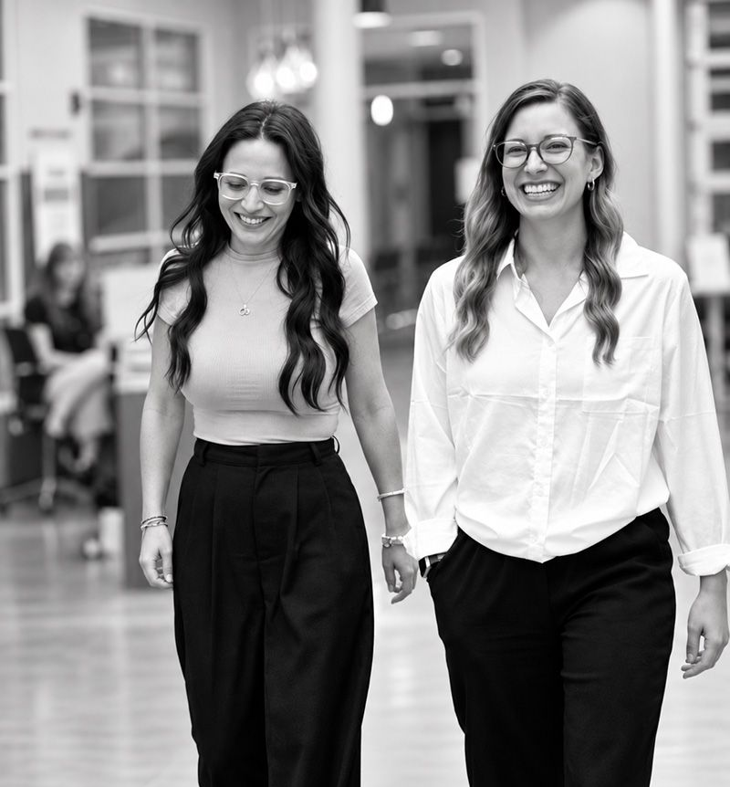Two smiling women walking together indoors, holding hands, one in white blouse and one in light top and dark pants