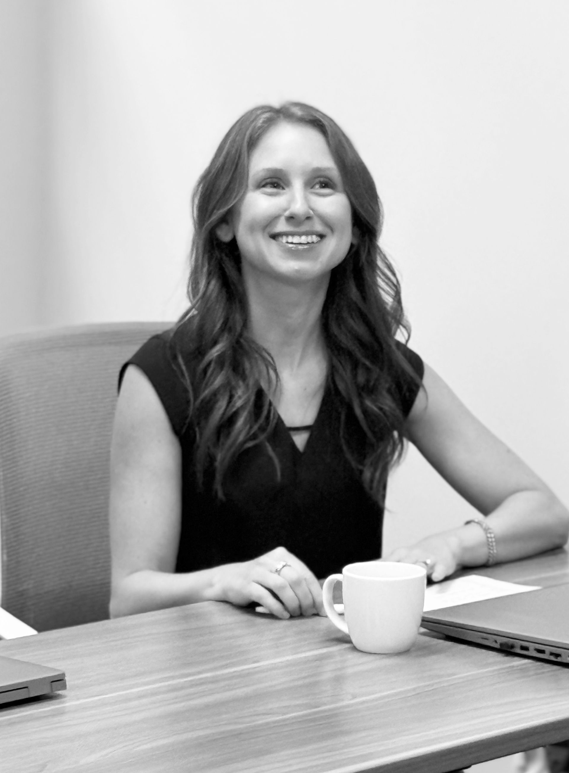 Smiling woman in a black top sitting at a table with a coffee mug and laptop in an office