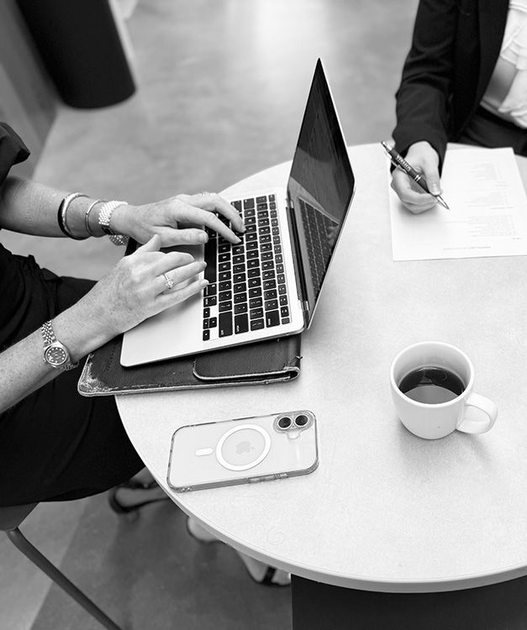 Hands typing on a laptop at a round table with coffee, notebook, and a phone on a white office desk.