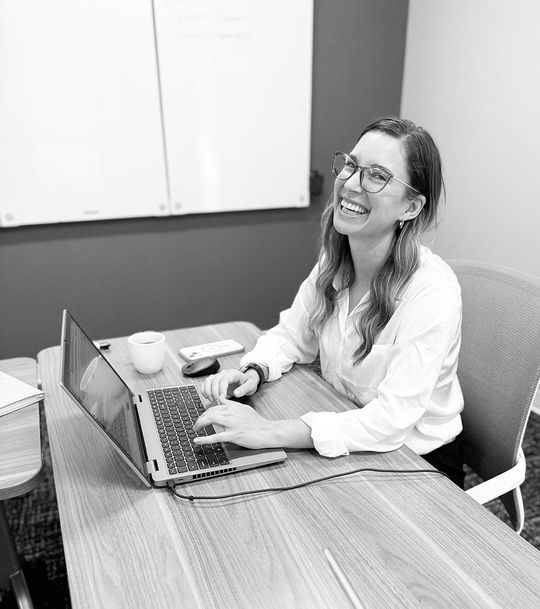 Woman smiling while typing on a laptop at a conference table in an office.