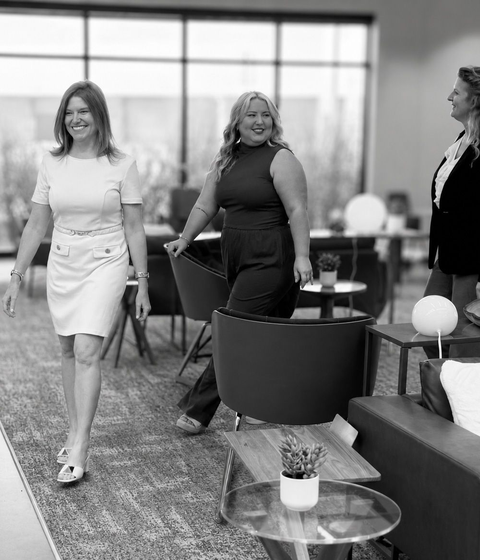 Two women walking through a modern office lounge, carrying chairs and smiling